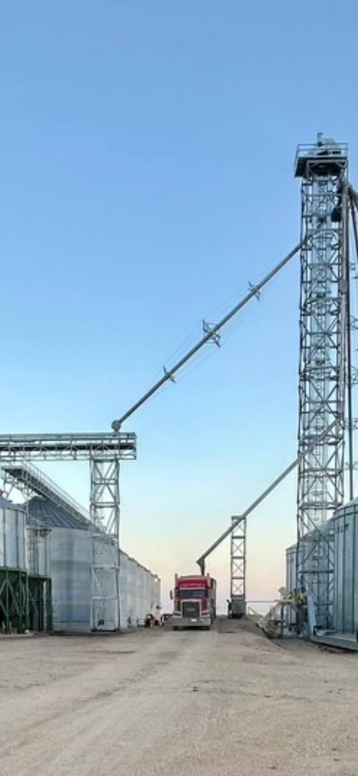 Galvanized RapiTower grain leg tower with spouting and a bridge conveyor over a bin yard, grain truck below